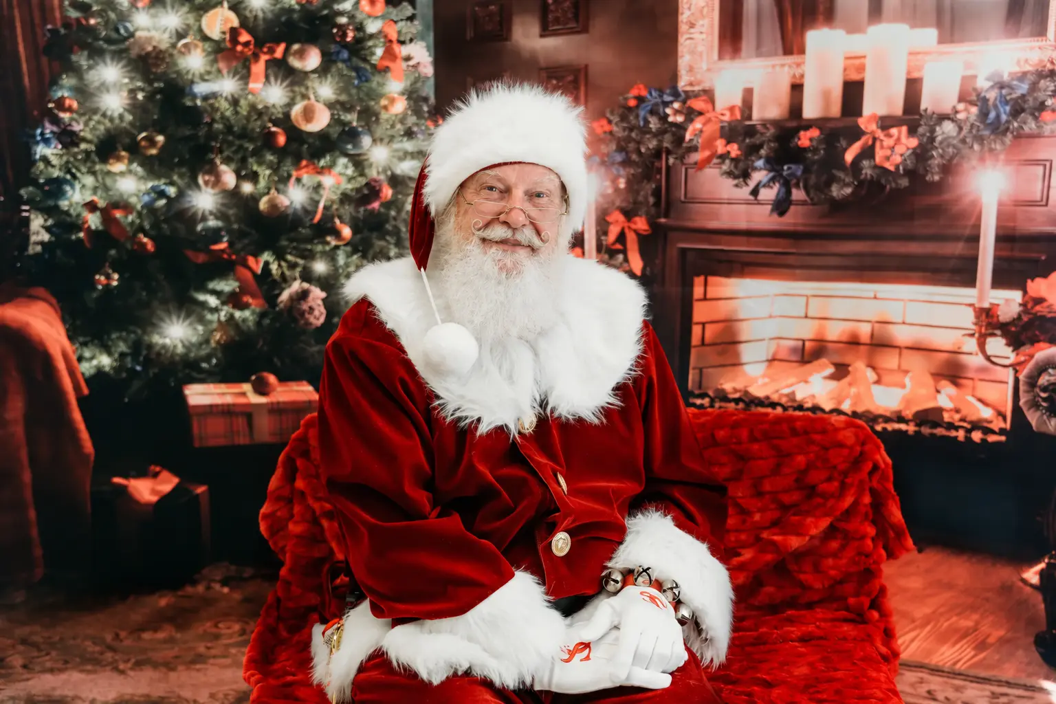Santa Skip seated beside a Christmas tree and fireplace
